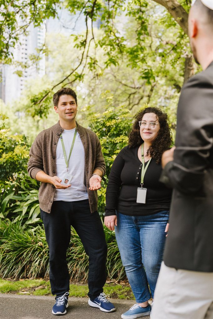 Three people wearing lanyards talk together on a path in a leafy park, with trees and city buildings blurred in the background.